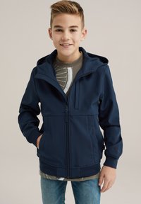 Mornarško modra zip-up kapuca s kapuco, z gladko teksturo, elastičnimi manšetami in sprednjim žepom, nosi se čez sivo grafično majico.