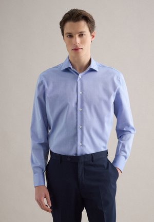 Junger Mann steht da, trägt ein hellblaues Hemd mit Knöpfen, das in dunkelblaue Hosen gesteckt ist, eine Hand in der Tasche, neutraler Hintergrund.