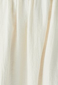 Un tessuto di colore crema dalla trama morbida si drappeggia delicatamente, mostrando morbide pieghe e una leggera lucentezza naturale. L'aspetto complessivo è etereo e delicato.