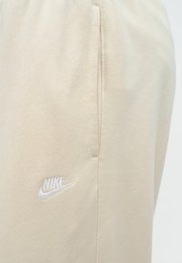 Calças de fato de treino bege da Nike com tecido texturizado, cintura elástica, bolsos laterais e um logótipo branco bordado na frente.