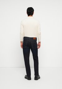 Tmavé denimové džiny s koženou nášivkou na zadnej strane, nosené s krémovou košeľou s dlhým rukávom; čierne topánky dopĺňajú outfit.