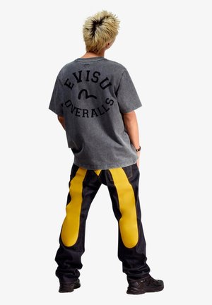 Persona con capelli corti biondi che indossa una maglietta grigia oversize con testo nero e pantaloni scuri con strisce gialle audaci dietro.