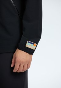 Black long-sleeve jacket featuring a zipper on the chest, with a fabric tag on the cuff displaying a multicolored mountain and sun design.