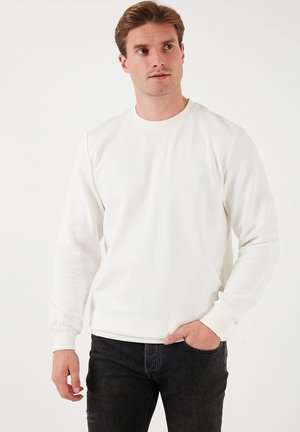 Witte sweatshirt met ribbelstructuur, ronde halslijn en lange mouwen. Gecombineerd met donkerere spijkerbroek, handen in de zakken.