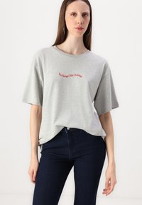 T-shirt gris à manches courtes avec un texte rouge indiquant "Sois sauvage, pas moyen." Associé à un jean bleu foncé, présenté sur un fond uni.