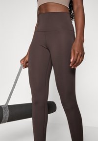 Brune højtaljede leggings lavet af strækbart materiale med en glat tekstur; hånd holder en rullet sort yogamåtte med en mønstret rem.