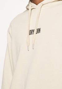 Beżowa bluza z kapturem wykonana z miękkiego materiału z troczkami zakończonymi srebrnymi końcówkami. Na wysokości klatki piersiowej wyhaftowany jest czarny napis "DRY BORN".
