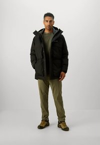 Crna podstavljena jakna s kapuljačom, dva prednja džepa i zatvarač, nošena preko zelene košulje i bež hlača, uparena s smeđim cipelama.