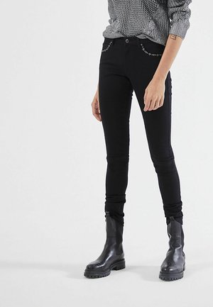 Personne portant un pantalon skinny noir clouté, un haut gris à motifs et des bottes en cuir noir épaisses, se tenant devant un fond uni.