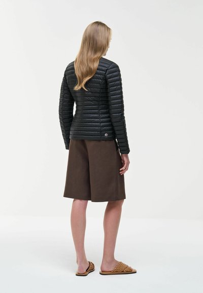 Femme aux longs cheveux blonds portant une veste matelassée noire, un short marron jusqu'aux genoux et des sandales tissées beiges, debout devant un fond uni.