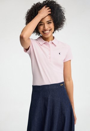 Femme souriante aux cheveux bouclés portant un polo rose clair et une jupe bleu foncé, la main touchant ses cheveux, debout devant un fond uni.