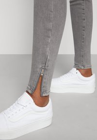 Vaqueros skinny grises con detalles de cremallera en el tobillo, combinados con zapatillas blancas. Material de denim suave con una textura lisa.