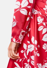 Rotes Blumenkleid mit weißen Blattmustern, seidiger Textur und langen Ärmeln. Die Manschetten sind mit dekorativen Knöpfen verziert, um zusätzliche Details zu bieten.