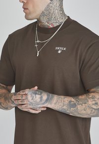 Camiseta de algodón marrón con mangas cortas, que presenta un logotipo blanco en el pecho. Modelo lleva collares de plata en capas, con los brazos cubiertos de tatuajes intrincados.