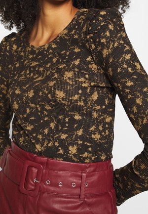 Femme portant un haut à manches longues noir et marron à taches avec des épaules bouffantes, rentré dans un pantalon en cuir rouge avec une large ceinture.