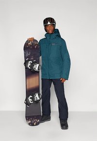 Teal vattentät jacka, mörka byxor, snowboardpjäxor; håller en snowboard med mörkt grafiskt mönster och vita bindningar.