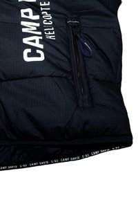 Chaqueta acolchada azul marino con patrón de acolchado horizontal, que presenta un bolsillo con cremallera en el frente, un logo de texto en blanco y una etiqueta interior negra.