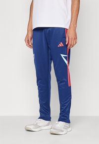 Vīrietis, kurš valkā zilas Adidas sporta bikses ar sarkaniem un gaiši ziliem ģeometriskiem akcentiem un baltām sporta kurpēm, stāvot pret vienkāršu fonu.