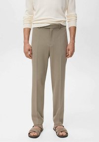 Mand iført beige skræddersyede bukser, cremet ribbet langærmet sweater og matchende beige sandaler med åben tå, står foran en hvid baggrund.