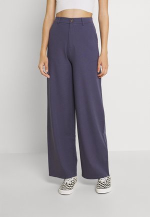 Person trägt hoch taillierte, weit geschnittene dunkelblaue Hose mit Knopfverschluss, weißes Crop-Top und schwarz-weiße karierte Sneakers.