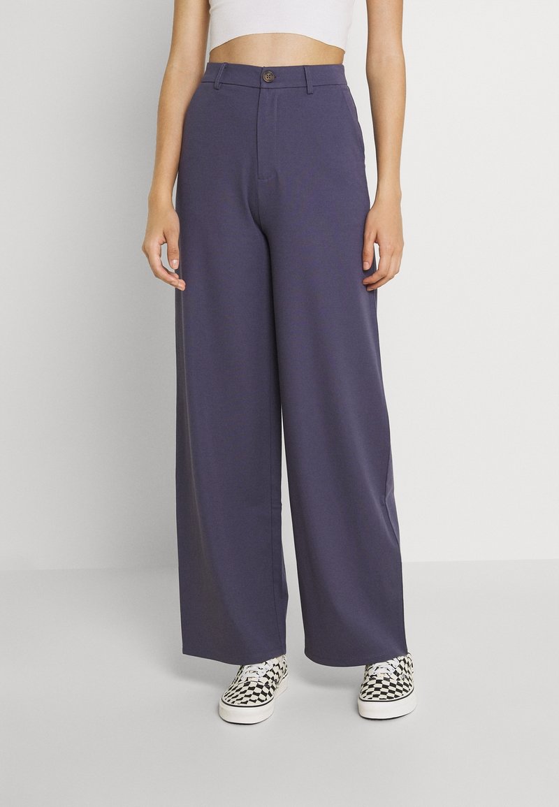 Personne portant un pantalon bleu foncé taille haute à jambes larges avec fermeture par bouton, un crop top blanc et des baskets à carreaux noir et blanc.