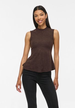 Femme souriante portant un haut sans manches marron foncé ajusté avec un léger évasement à l’ourlet et un jean noir slim devant un fond blanc.