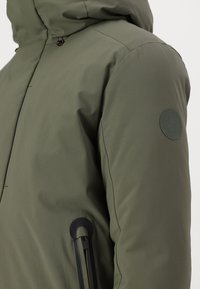 Olivegrüne Kapuzenjacke mit glatter Textur, ausgestattet mit einem Gummi-Logopatch am Oberarm und kontrastierenden schwarzen Reißverschlüssen.