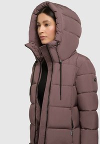 Jachetă căptușită în nuanța mauve, cu o glugă oversized, fermoare negre și accente cu șnur. Materialul are o textură netedă, cu secțiuni pătrate matlasate.