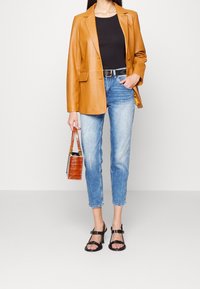 Woman wearing a tan leather blazer, black top, light blue jeans, black sandals, and holding a small brown handbag with crocodile texture.