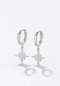 Srebrne kolczyki w kształcie kółek mają zawieszkę w kształcie gwiazdy, ozdobioną przezroczystymi kamieniami, które odbijają światło i rzucają wyraźne cienie.