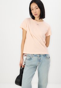 Tricou din bumbac roz deschis, cu mâneci scurte, decolteu rotund și un mic logo negru. Asortat cu blugi din denim deschis și o geantă neagră.