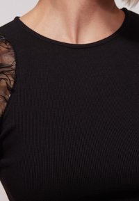 Černý žebrovaný top s průhlednou, strukturovanou látkou na ramenou, která zvýrazňuje kontrast mezi materiály a designovými prvky.