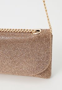 Metaliczna torebka w kolorze różowego złota z teksturowaną powierzchnią pokrytą małymi kryształkami, zamknięciem z złożonym klapą oraz złotym łańcuszkiem jako paskiem.