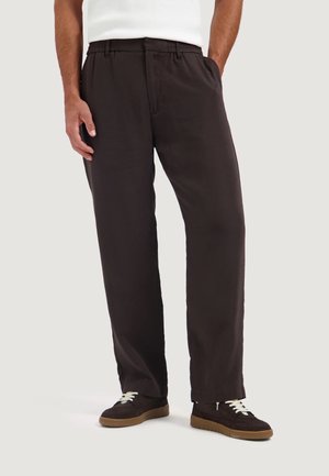 Hombre con pantalones negros holgados con bolsillos laterales y zapatillas negras con cordones blancos sobre un fondo liso.