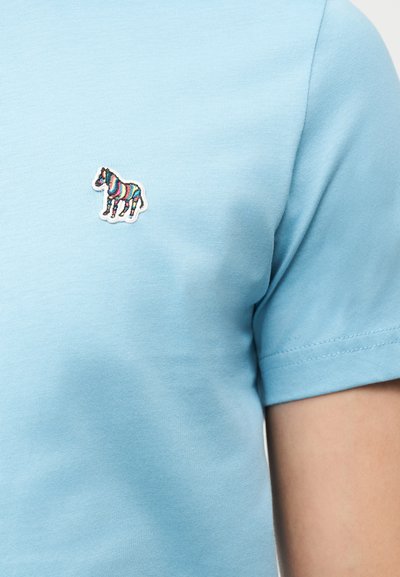 Ανοιχτό μπλε πουκάμισο με κοντά μανίκια και μικρό πολύχρωμο ριγέ έμβλημα ζέβρας στο στήθος.