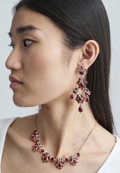 Rød smykkesett med edelstener som inkluderer lysekroneøredobber og en halskjede. Gullfargede detaljer med blomsterdesign, flere former og teksturer.