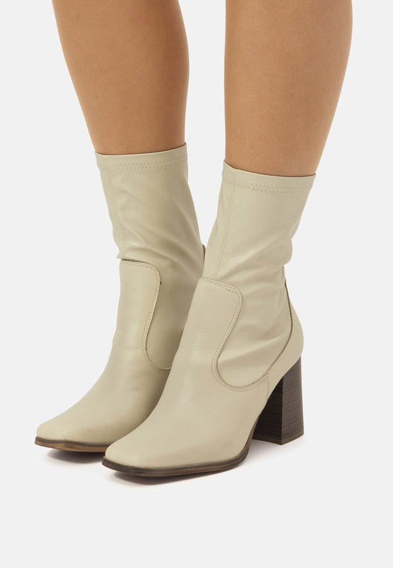 Botas de tobillo de cuero beige con punta cuadrada, caña elástica y tacón cuadrado de 7 cm. Presenta costuras sutiles y detalles recortados en los costados.