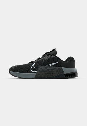 Chaussure de sport noire avec tige en maille, accents gris et semelle intermédiaire texturée. Présente un logo proéminent et un design à lacets. Semelle extérieure en caoutchouc pour une meilleure adhérence.