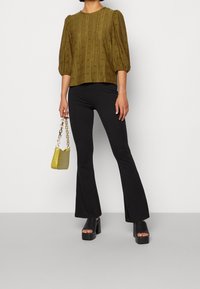 Top in verde oliva con texture e maniche a palloncino, abbinato a pantaloni neri a palazzo e scarpe platform nere robuste. Tiene in mano una piccola borsa gialla.
