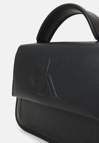 Svart läderhandväska med präglad cK-logotyp, strukturerad form, slät yta och ett topphandtag. Har en minimalisk design och rena linjer.