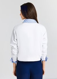 Felpa bianca con maniche lunghe, taglio corto, con una camicia colletto a righe blu e bianche indossata sotto. Texture liscia.