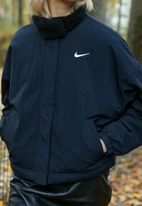 Mörkblå jacka med en plysch, svart krage, elastiska ärmslut och sidfickor. Har en vit Nike-logotyp på övre vänstra bröstet.