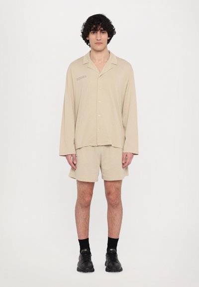 Beige košeľa s krátkym rukávom a uvoľneným strihom, v kombinácii s rovnakými béžovými šortkami. Čierne topánky a ponožky dodávajú outfitu celkový vzhľad.