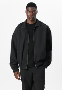 Schwarze Bomberjacke mit Reißverschluss, strukturierter Oberfläche, elastischen Bündchen und Saum. Mit subtiler Logodetails auf der Brust.