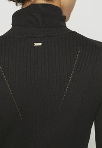 Czarny sweter z golfem w prążki, z teksturowanym wzorem, z odkrytymi plecami i małą złotą metką przy dekolcie.