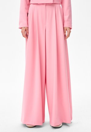 Persoon die roze, hoogwaardige, wijduitlopende broek draagt met een bijpassend roze jasje, staand tegen een effen witte achtergrond.