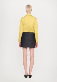 Camicia gialla a maniche lunghe, mini gonna in denim scuro, vista posteriore. Vestibilità slim, texture liscia, dettaglio di cucitura sottile, scarpe slip-on nere.