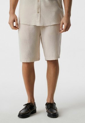 Mężczyzna ubrany w beżowe spodenki do kolan, dopasowaną zapinaną koszulę oraz czarne skórzane buty sznurowane, stojący na jasnym, prostym tle.