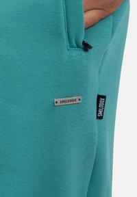 Turkusowy sweatshirt o gładkiej fakturze, z boczną kieszenią i markowymi metkami. W zestawie znajduje się mała metalowa etykieta z napisem "SMILODOX."