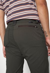 Mörk olivgröna byxor med en dragkedjeförsedd bakficka, subtila sömnadsdetaljer och en märkeslapp på midjebandet.